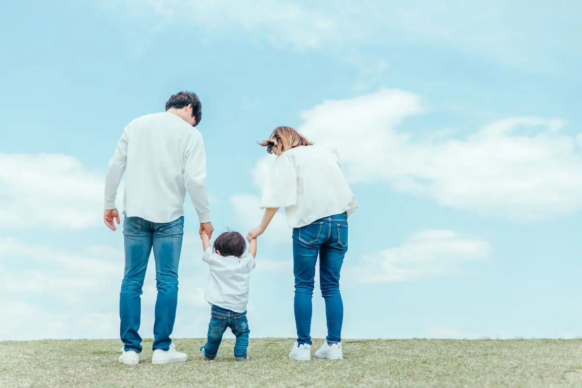 青空の下、家族3人が手をつないで歩く後ろ姿(子育て支援住宅のイメージ)